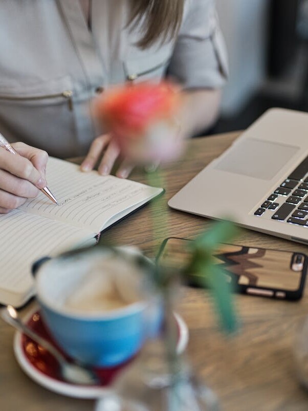 Person Writing On A Notebook Beside Macbook