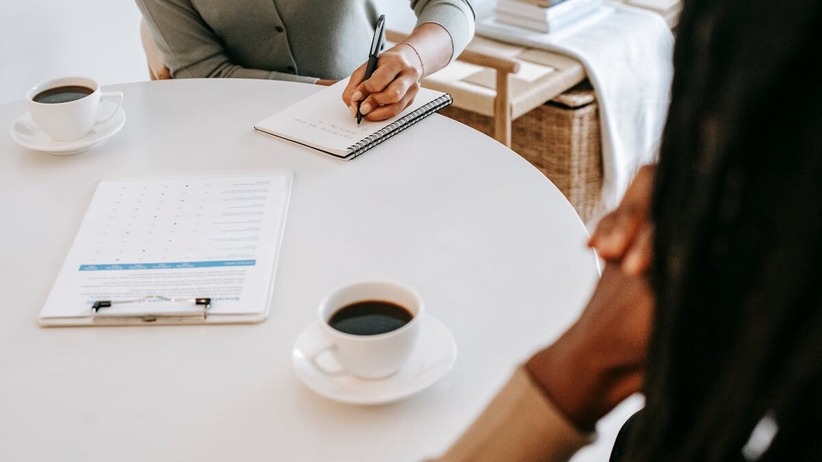 From above crop multiracial female interviewer or psychologist in formal wear asking questions and taking notes in planner while talking to black man and sitting together at table with coffee