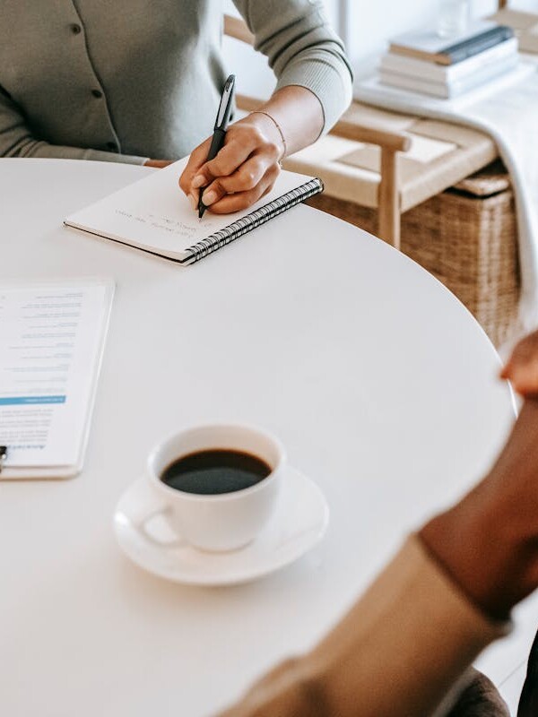 From above crop multiracial female interviewer or psychologist in formal wear asking questions and taking notes in planner while talking to black man and sitting together at table with coffee