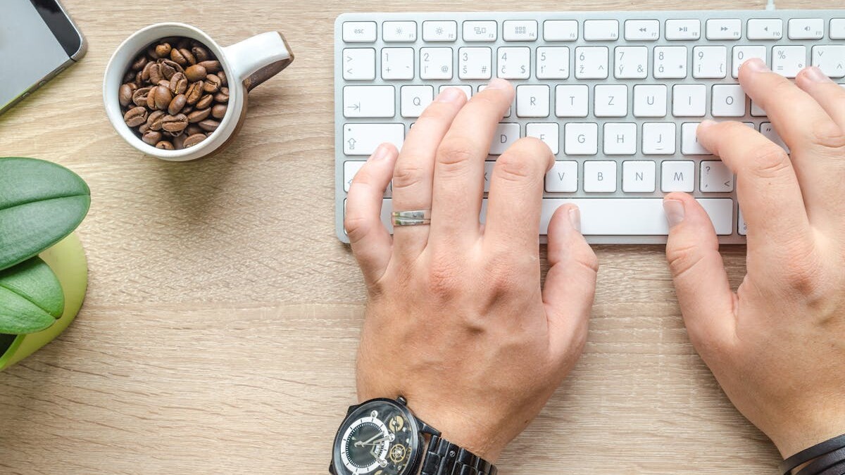 Person Using Silver Apple Magic Keyboard Beside of White Ceramic Mug With Coffee Beans