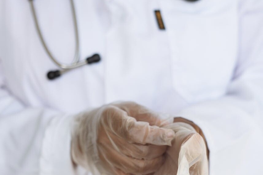 Faceless ethnic medical worker in lab coat and stethoscope taking of transparent gloves after approaching patients for examination in modern hospital