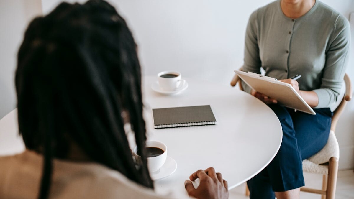 Professional ethnic female psychotherapist asking questions to black male patient and taking notes in clipboard while sitting together at table in modern psychotherapy center