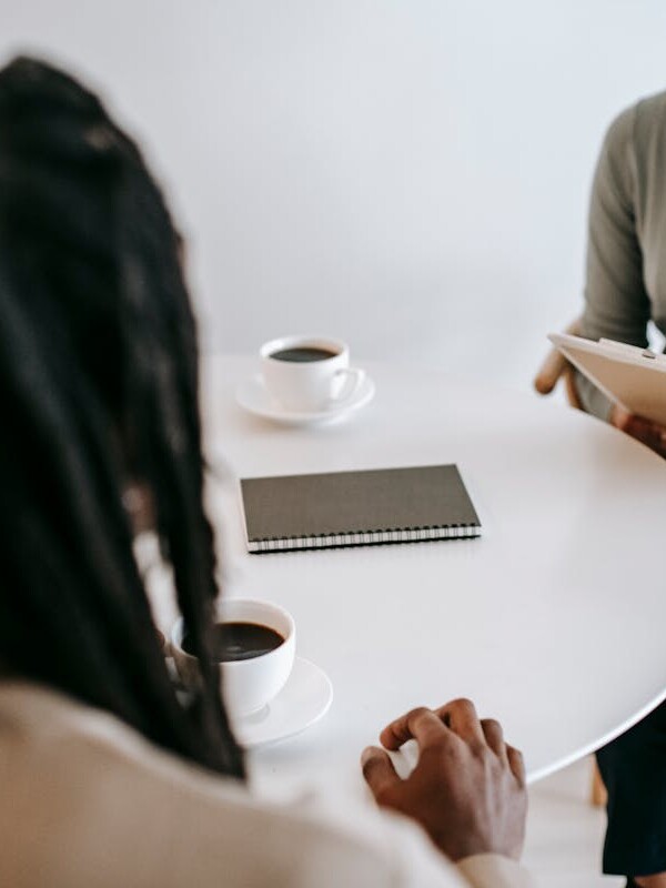 Professional ethnic female psychotherapist asking questions to black male patient and taking notes in clipboard while sitting together at table in modern psychotherapy center