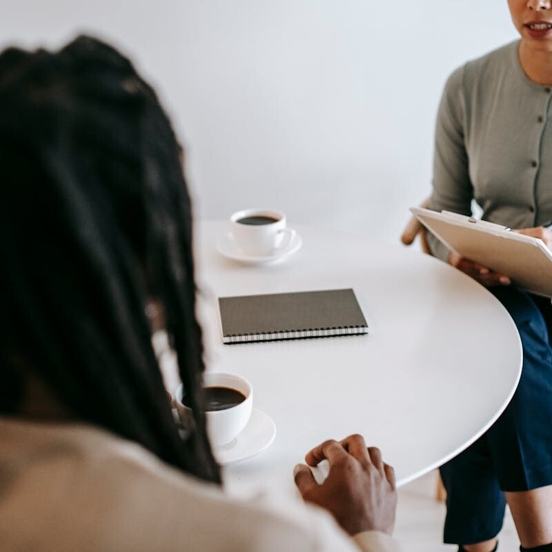 Professional ethnic female psychotherapist asking questions to black male patient and taking notes in clipboard while sitting together at table in modern psychotherapy center