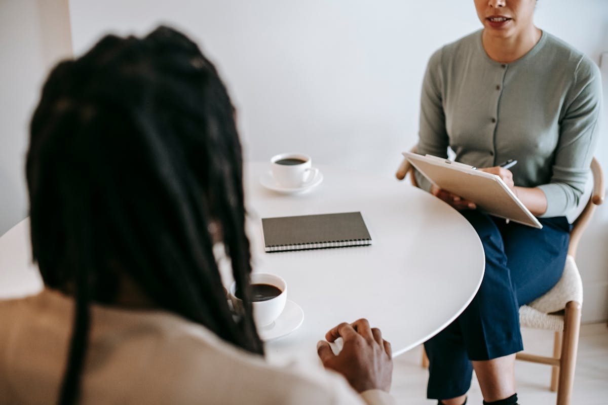 Professional ethnic female psychotherapist asking questions to black male patient and taking notes in clipboard while sitting together at table in modern psychotherapy center