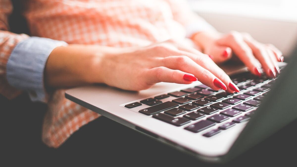 Close-up of a woman with red nails typing on a laptop indoors.
