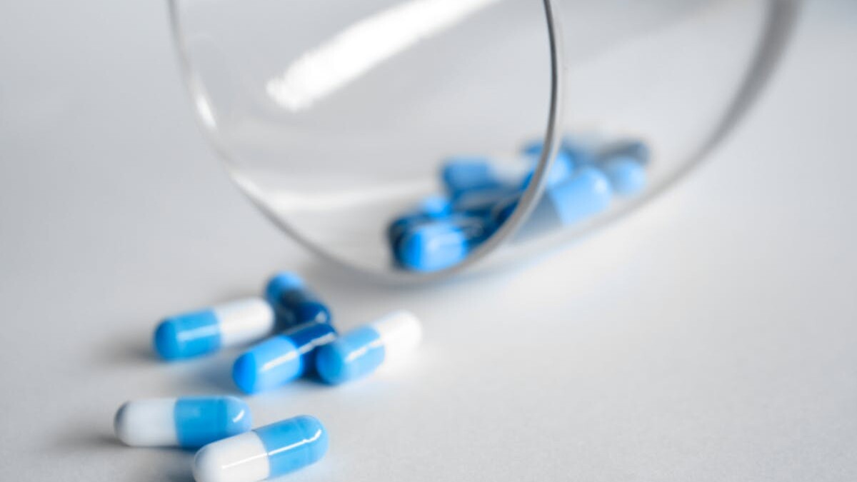 A close-up of blue and white capsules spilling from a glass on a light background.