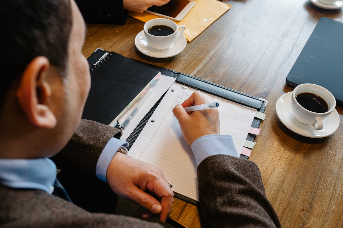 A professional man writes notes at a meeting table with coffee cups.