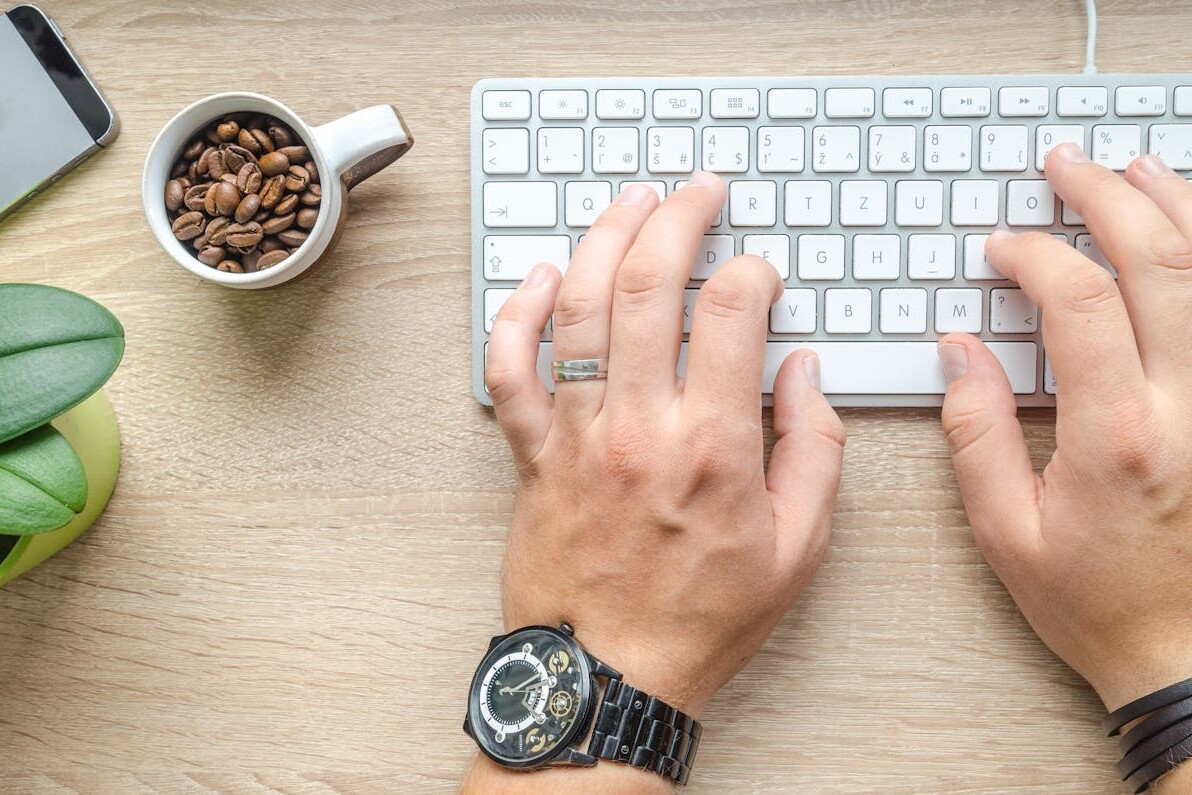 Hands typing on a keyboard beside a cup of coffee and a smartphone on a wooden table.