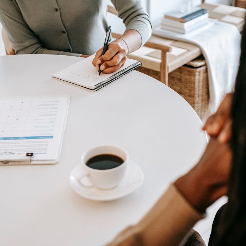 From above crop multiracial female interviewer or psychologist in formal wear asking questions and taking notes in planner while talking to black man and sitting together at table with coffee