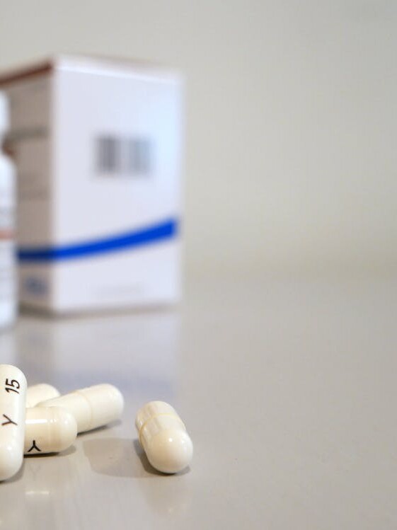 Close-up image of white capsules with medicine bottle and box in the background.